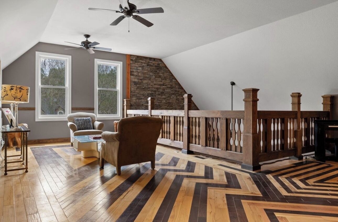 A spacious loft with a high ceiling, exposed wooden beams, a large window, and a chevron-patterned floor.