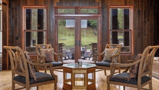 Una habitación con paredes de madera y ventanas amplias, dos sillas de madera con cojines, y una mesa con superficie de vidrio.
