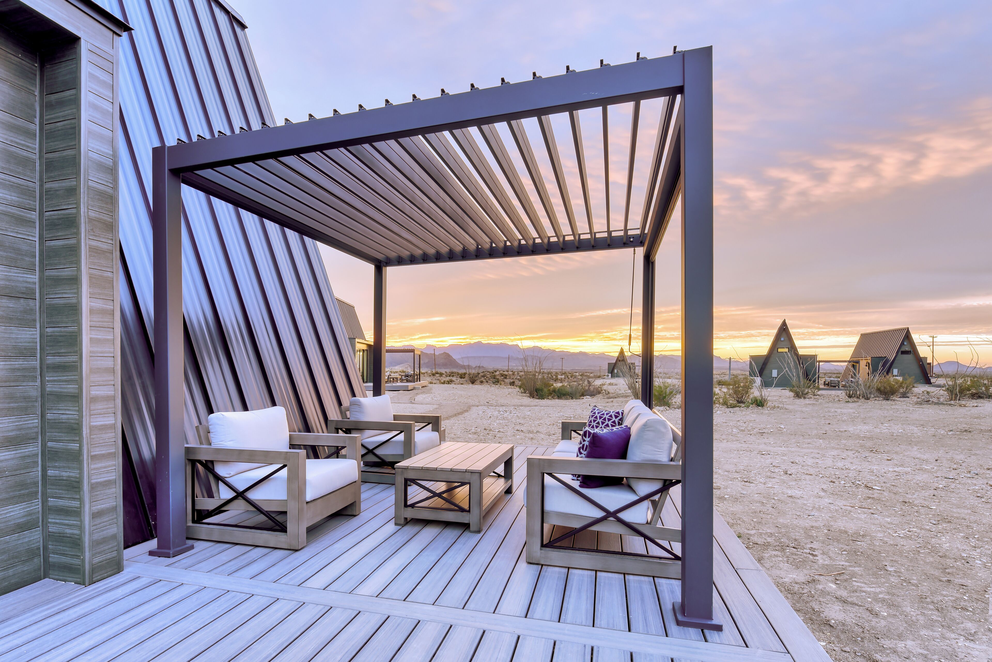 Une terrasse en bois avec des sièges d’extérieur, une pergola et une vue sur le paysage désertique au coucher du soleil.