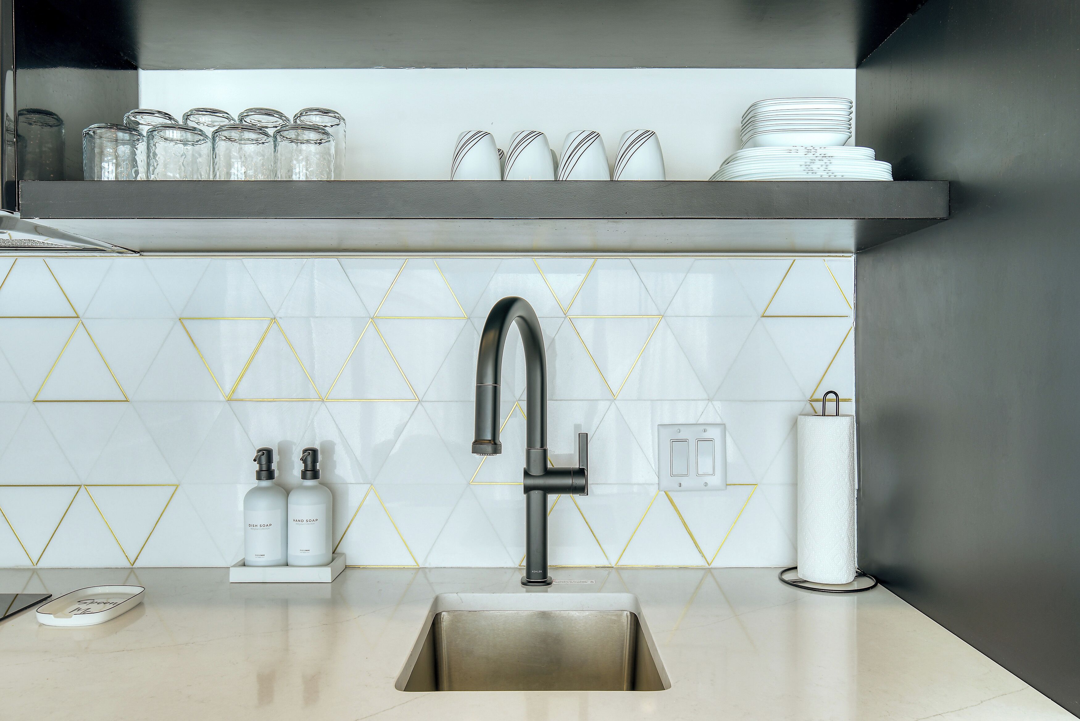 A modern kitchen with a stainless steel sink, a faucet, and a shelf above holding glassware and plates.