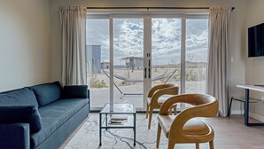 Un salon moderne avec un canapé, une table basse en verre et deux fauteuils. Une grande porte-fenêtre coulissante donne accès à un balcon équipé d’un hamac et offrant une vue sur la plage.