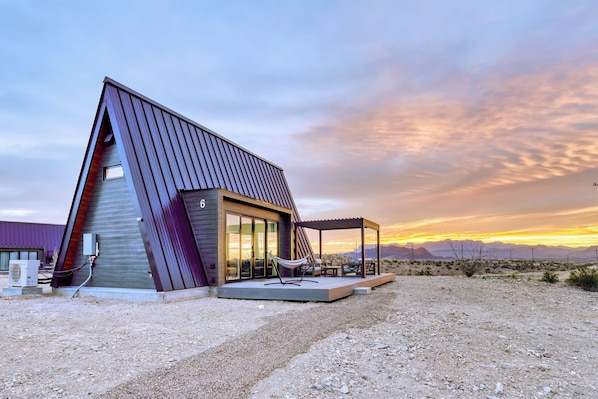 A modern house with a sloped roof, a covered patio with outdoor seating, and a desert landscape in the background. - Stardust Big Bend Luxury A-Frames (Terlingua)