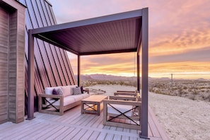 A covered outdoor seating area with a sofa and chairs, overlooking a desert landscape at sunset. - Stardust Big Bend Luxury A-Frames (Terlingua)