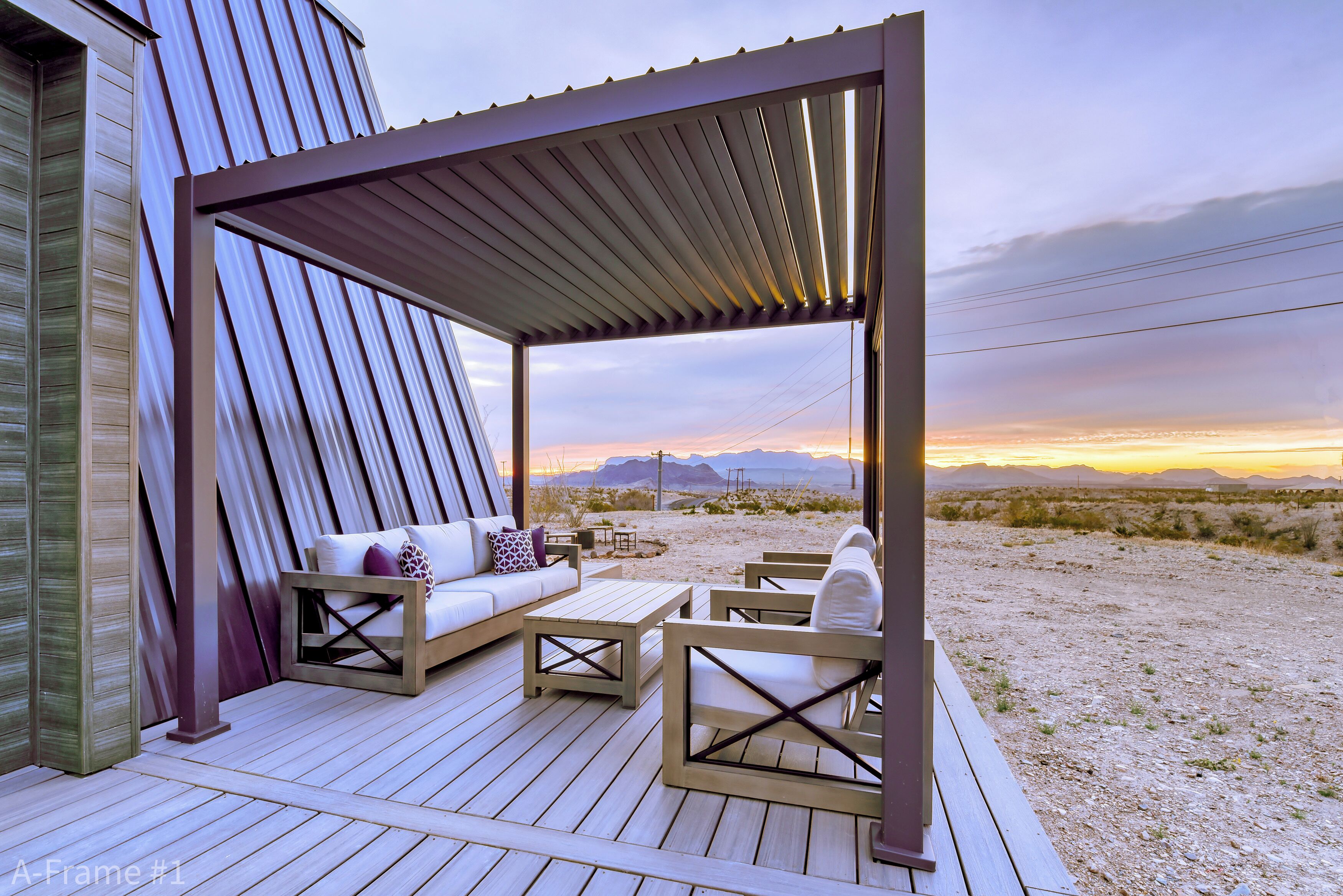 A covered outdoor seating area with a sofa and chairs, a wooden table, and a view of a desert landscape at sunset.