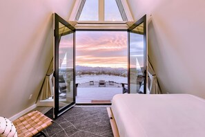A bedroom with a large bed, a bench, and a view of a snowy landscape through a glass door. - Stardust Big Bend Luxury A-Frames (Terlingua)