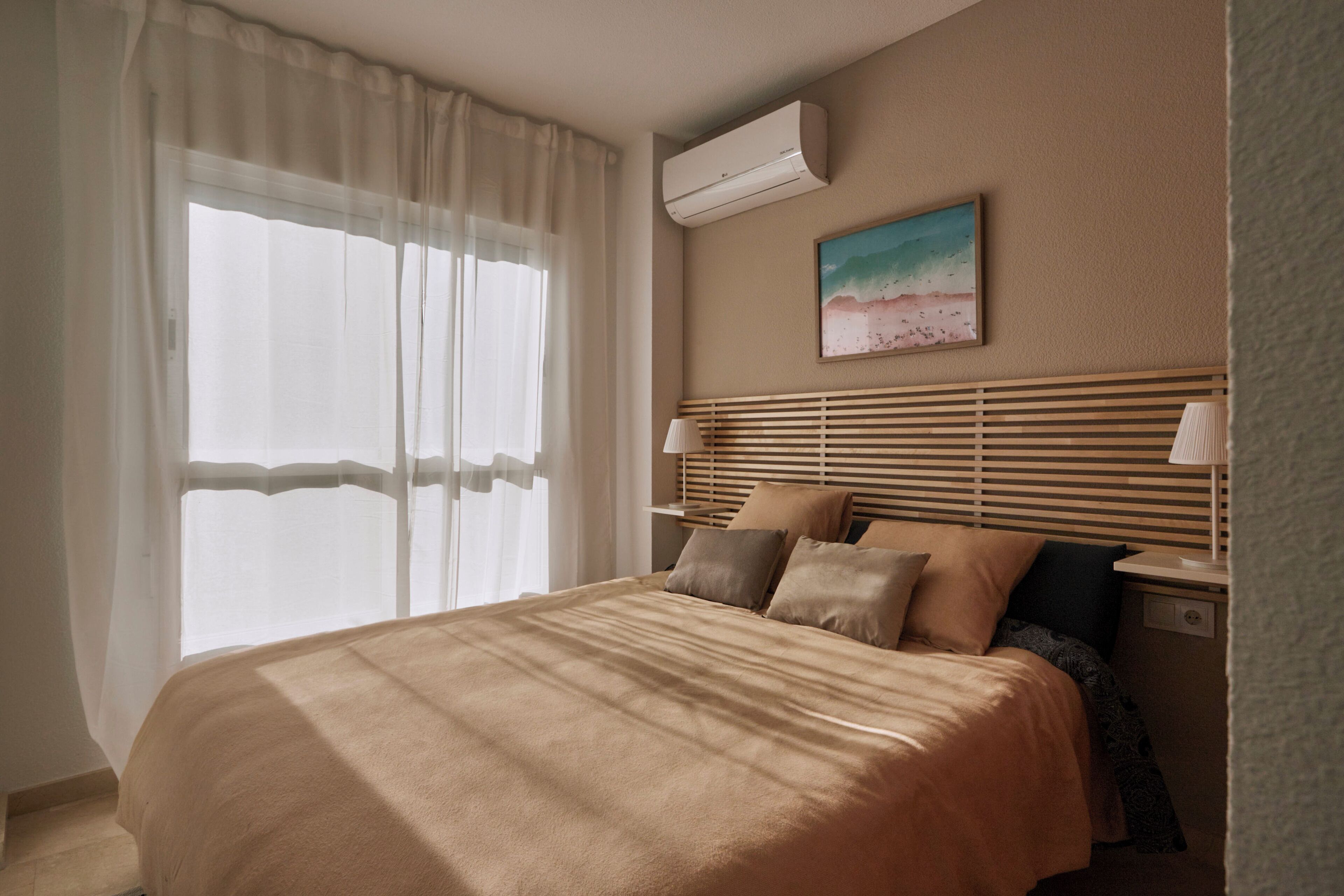 A neatly arranged hotel room with a bed, a bedside table with a lamp, a wall-mounted air conditioner, and a beach-themed painting.
