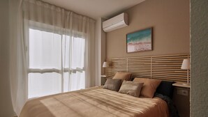 Une chambre d’hôtel bien aménagée, avec un lit, une table de chevet dotée d’une lampe, un climatiseur fixé au mur et un tableau sur le thème de la plage.