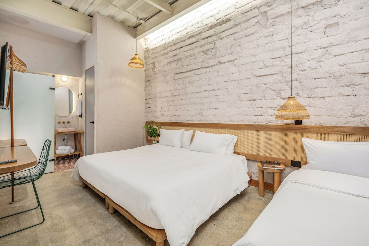 A modern hotel room with a large bed, a wooden nightstand, a small stool, a desk with a chair, and a bathroom with a mirror and sink.