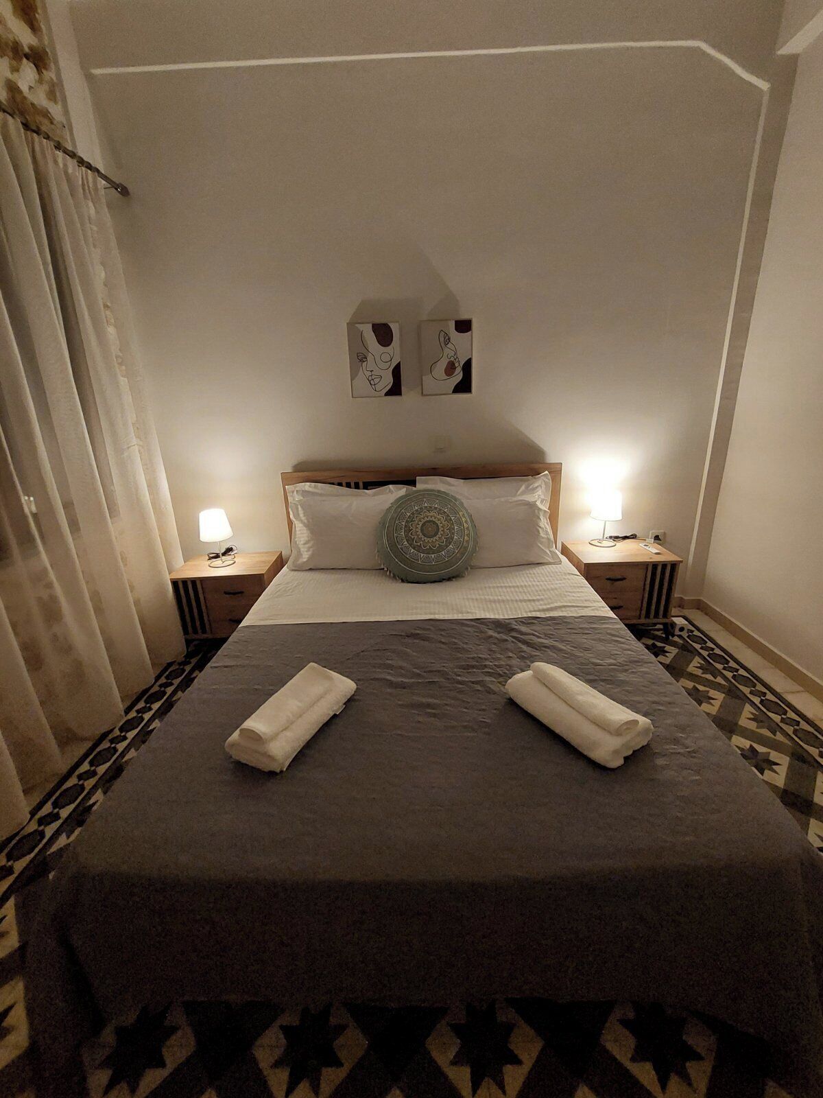 Un lit bien fait, recouvert d’une couvre-lit grise, avec deux serviettes blanches et un coussin décoratif. On y trouve deux tables de chevet avec des lampes, un tapis à motifs et un rideau sur le côté gauche.