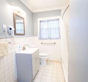 Ein Badezimmer mit weißem Waschbecken, WC und Dusche. Ein Fenster mit gemustertem Vorhang, und ein Fön an der Wand.