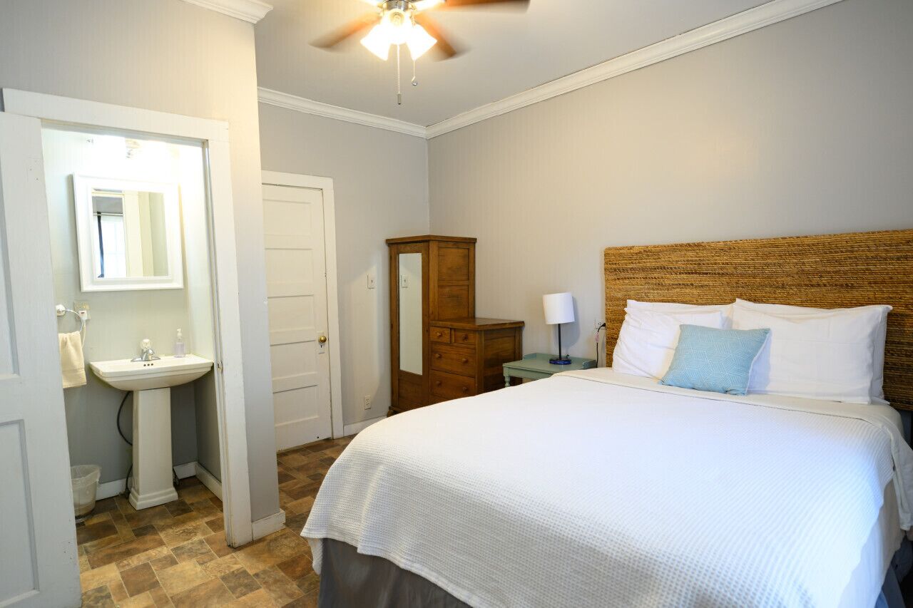 Un dormitorio con cama, cabecero de madera, mesita de noche con lámpara, cómoda y baño con lavabo y espejo.