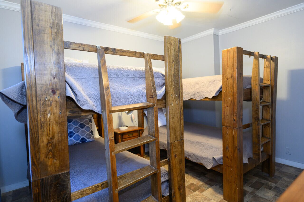 Quarto com duas beliches de madeira, uma pequena mesa de cabeceira e um abanador de teto.