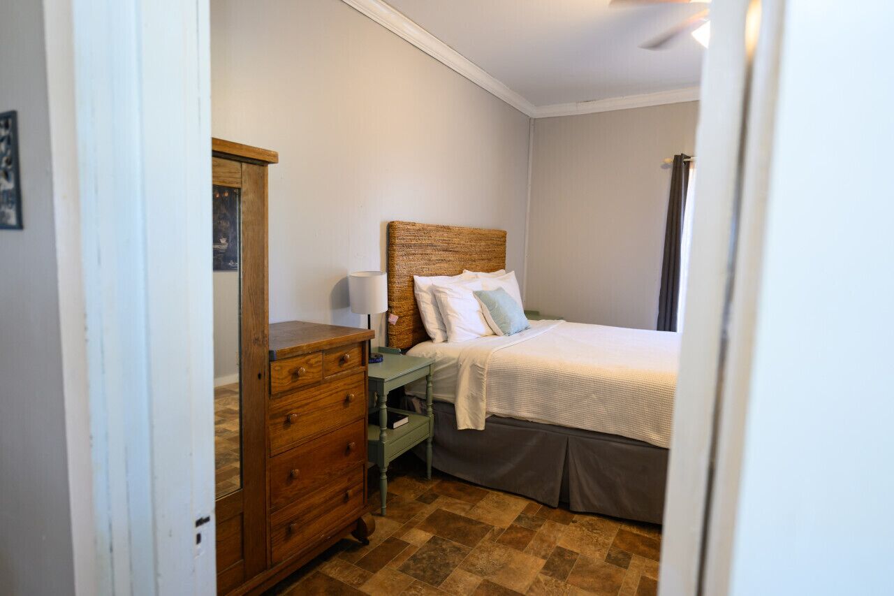 Un dormitorio con cómoda de madera, mesita de noche con lámpara, cama con cabecera y ventana con cortinas.