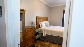 Un dormitorio con cómoda de madera, mesita de noche con lámpara, cama con cabecera y ventana con cortinas.