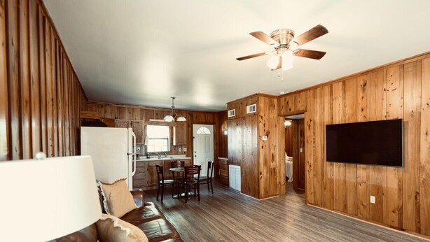 Un vaste espace de vie doté d’un plancher en bois, d’un ventilateur de plafond, d’un téléviseur à écran plat fixé au mur, et d’un coin repas avec des chaises et une table.