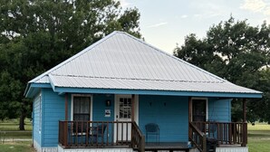 Un petit bâtiment d’un seul étage, à l’extérieur bleu et doté d’un toit en métal, avec une véranda ornée de balustrades en bois et un petit escalier menant à l’entrée.