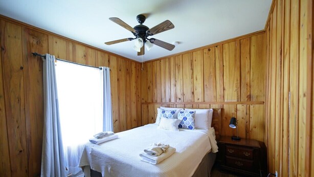 A bedroom with a bed, a nightstand, a ceiling fan, and wooden walls.