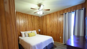 Een slaapkamer met een bed, plafondventilator, houten wanden, een raam met gordijnen en een televisie.