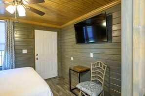 Una habitación con cama, una silla, televisión montada en la pared y ventilador de techo.