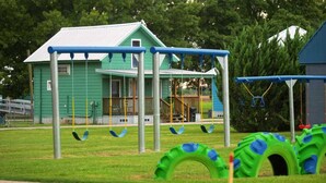 Un parque infantil con columpios y un neumático colgante, un edificio verde al fondo y una zona cubierta de césped.