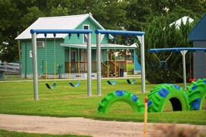 Un parque infantil con columpios y un neumático colgante, un edificio verde al fondo y una zona cubierta de césped.
