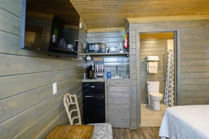 A compact kitchen with a refrigerator, coffee maker, and sink. A small dining table and chair are visible. The room has wooden walls and a wooden ceiling. - Serene cabin, relax and recharge! Great Internet! (Lake Village)