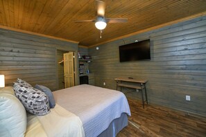 A bedroom with a bed, pillows, a ceiling fan, a TV, and a small kitchenette.