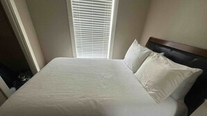 Una cama bien hecha con cubrecama y almohadas blancas, un cabecero de madera y una ventana con persianas.