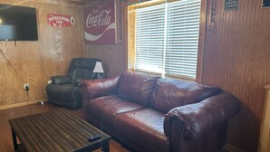 Een kamer met een leren bank, een aan de muur bevestigde televisie, een Coca-Cola bord en een raam met luxaflex.