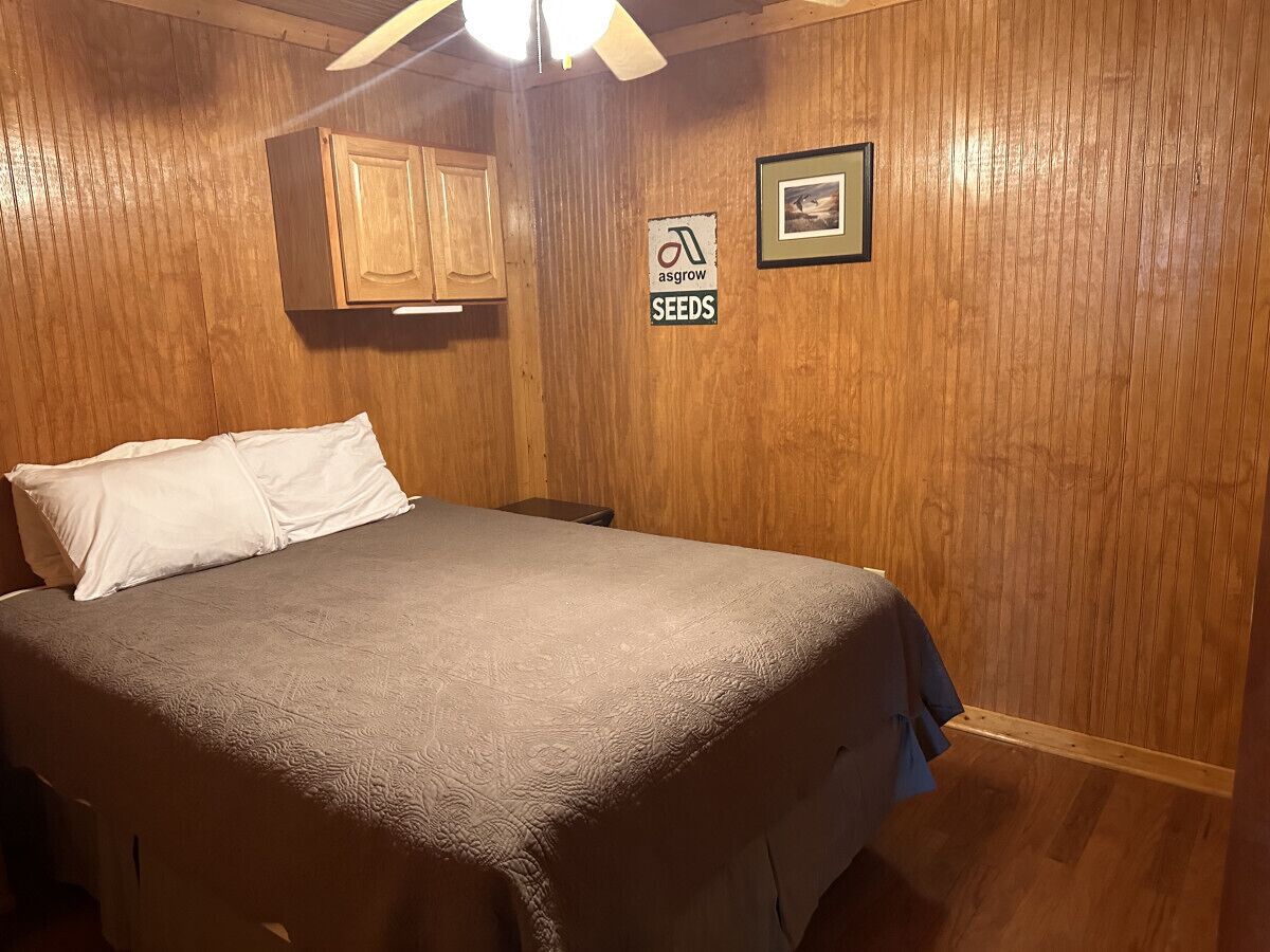 Een slaapkamer met een bed, houten wanden, een plafondventilator, een bord met 'asgrow SEEDS' en een lijst met een afbeelding.