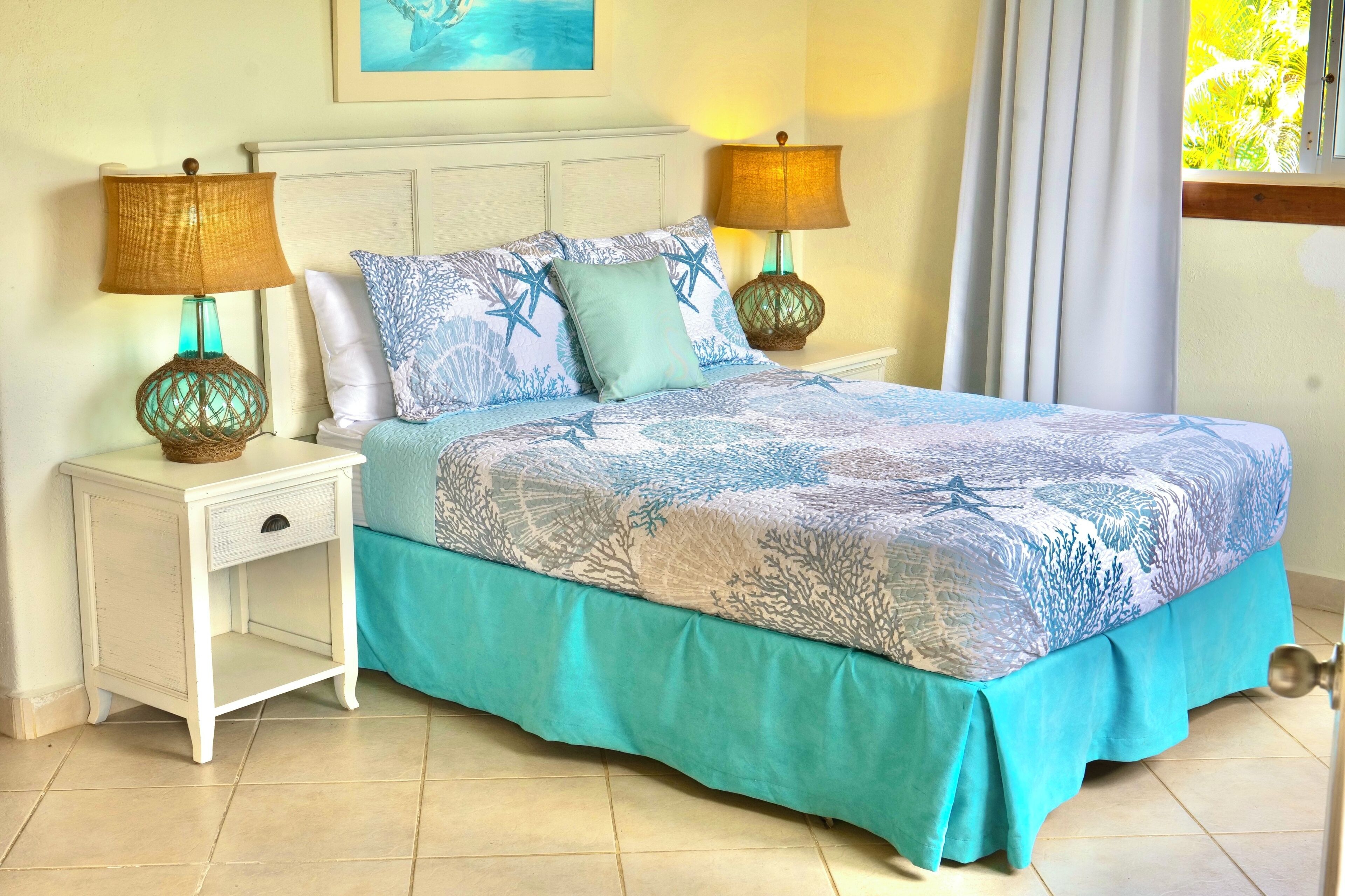 Cama bem arrumada com colcha azul e branca em padrão de corais, duas luminárias iguais e uma mesa de cabeceira com gaveta.