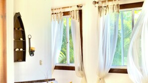 A room with a white sofa, a small white table, and a wicker basket. There are two windows with curtains, and a ceiling fan is visible.