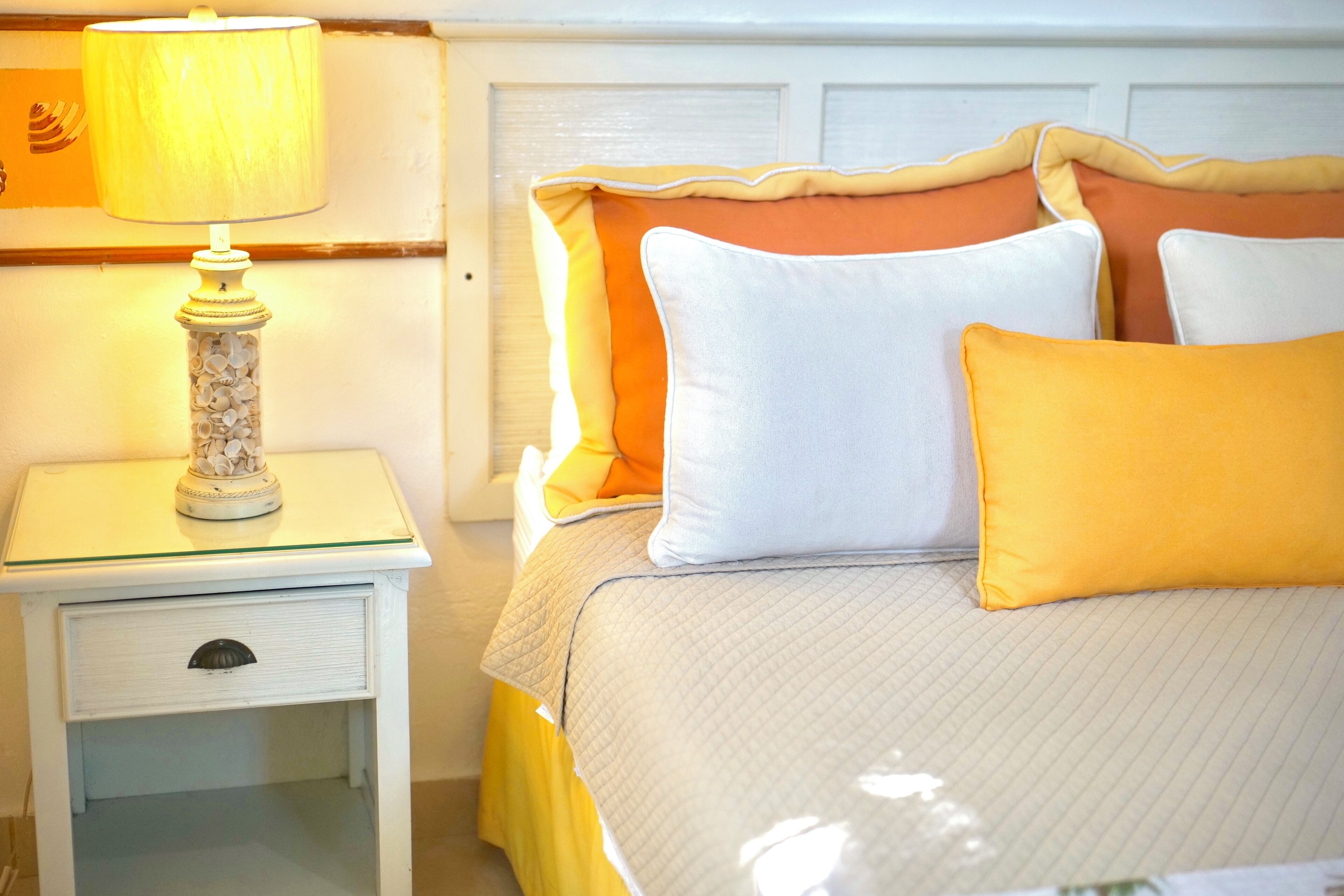A neatly made bed with a quilted cover, a bedside lamp, and a small white nightstand with a drawer.