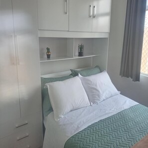 A bedroom with a bed, white and green bedding, a wardrobe with drawers, and a shelf with decorative items. - Apartamento Funcional em Curitiba (Curitiba)