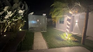 Un espacio exterior nocturno con un sendero, una zona de estar y un pequeño jardín con plantas e iluminación.