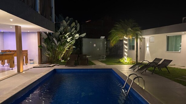 Una piscina con sillones de descanso, una mesa y un pequeño jardín con plantas.
