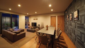 A modern living room with a large sofa, dining table, and chairs. There is a TV and a small table with decorative items. The room has large windows and a view of the outside. - Sanwa sen (Beppu)