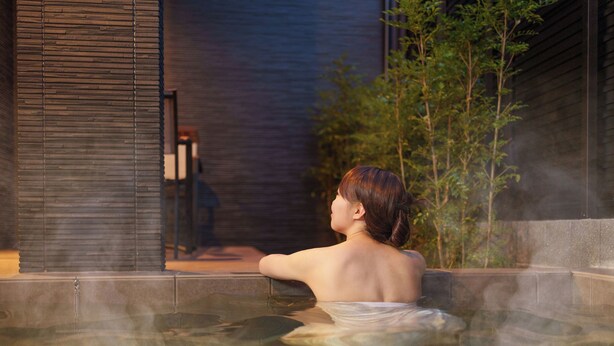 A person in a hot spring with a view of a building and greenery. - Sanwa sen (Beppu)