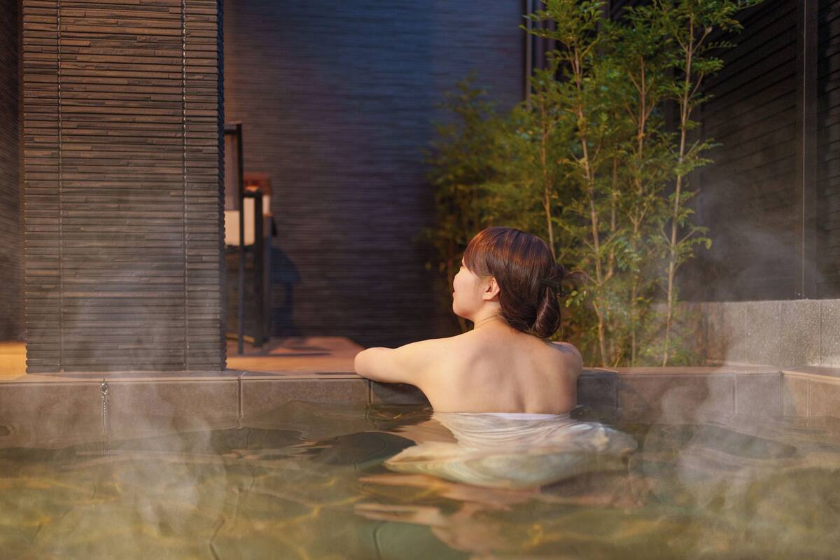 A person in a hot spring with a view of a building and greenery.