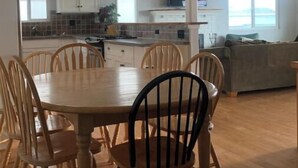 Une cuisine moderne avec un coin repas, comprenant une grande table et des chaises en bois, un éclairage suspendu et une vue sur la mer grâce à de grandes fenêtres.