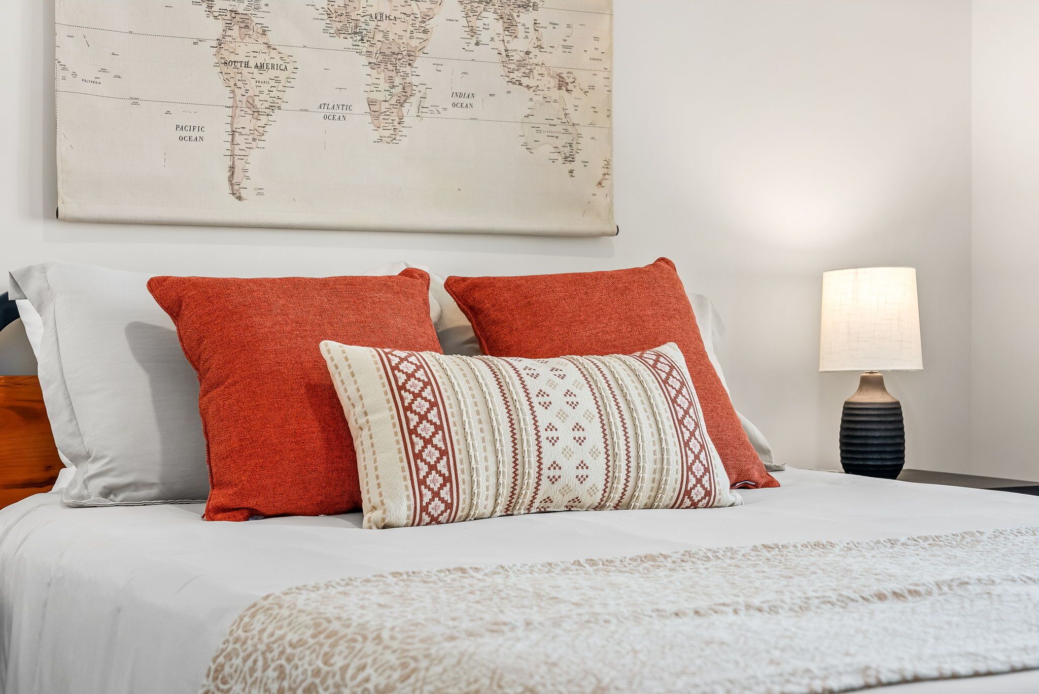 Un lit bien fait, avec deux oreillers rouges et un oreiller à motifs, une carte du monde accrochée au mur et une lampe sur la table de chevet.