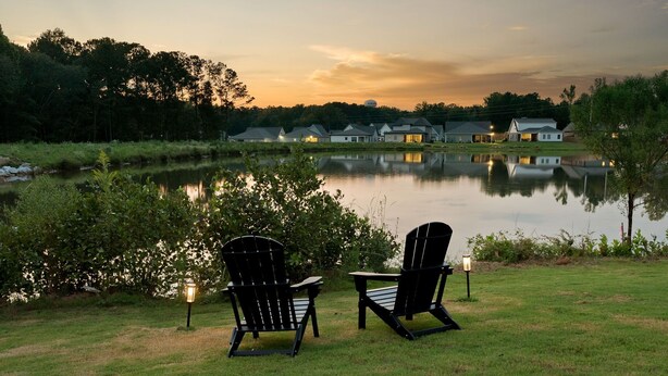 Dos sillas Adirondack junto a un lago tranquilo, con una fila de casas al fondo.