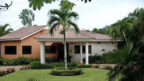 Una casa de dos pisos con techo de tejas, un porche cubierto y un jardín bien cuidado con palmeras y arbustos.