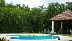 Una piscina con techado de azulejos y un porche, rodeada de exuberante vegetación y sillas de descanso.
