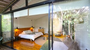 A modern bedroom with a large bed, a bedside table, and a nightstand. There is a window with a view of trees outside.