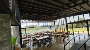 A spacious dining area with tables and chairs, large windows offering a view of a lake and hills, and a stone wall on one side.