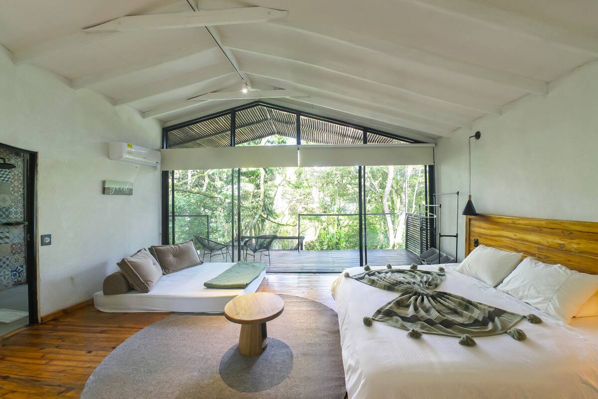 A modern bedroom with a large bed, a seating area with a sofa and chairs, and a view of greenery through large windows.