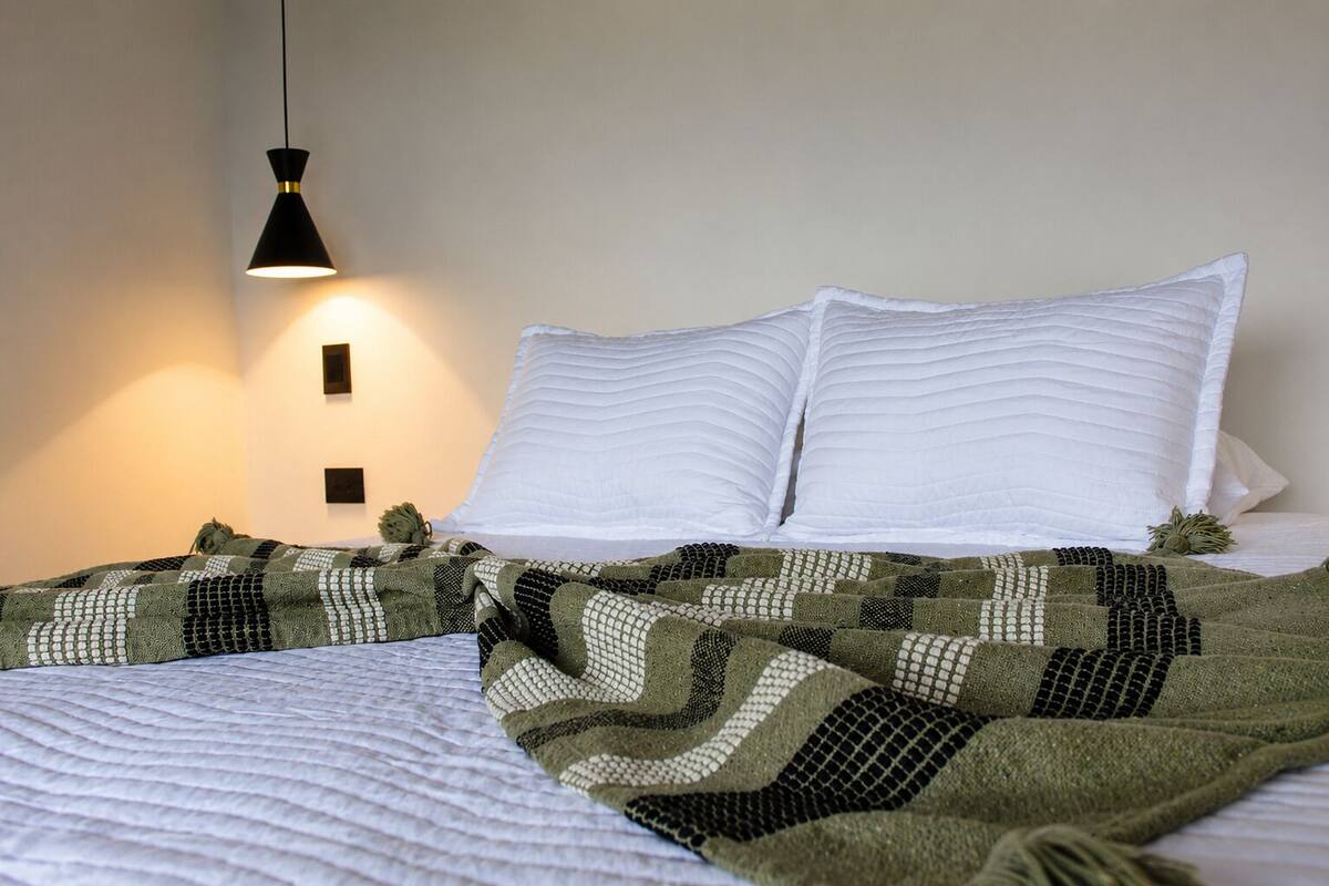 A bed with white pillows and a green and black patterned blanket, a black pendant light, and a wall-mounted light fixture.