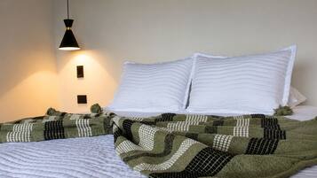 Una cama con almohadas blancas y una manta estampada en verde y negro, una lámpara colgante negra y un aplique de pared.