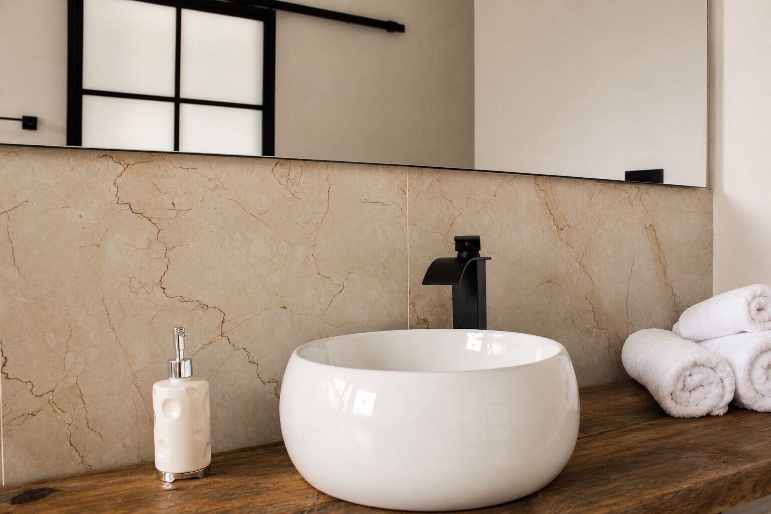 A bathroom with a large white vessel sink, a black faucet, a soap dispenser, and rolled towels on a wooden countertop.